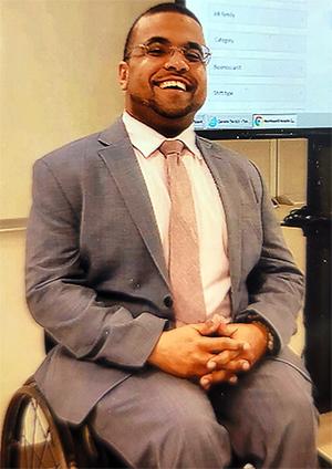 Lesly St. Louis is a black man that sits in his wheelchair wearing a suit and tie.