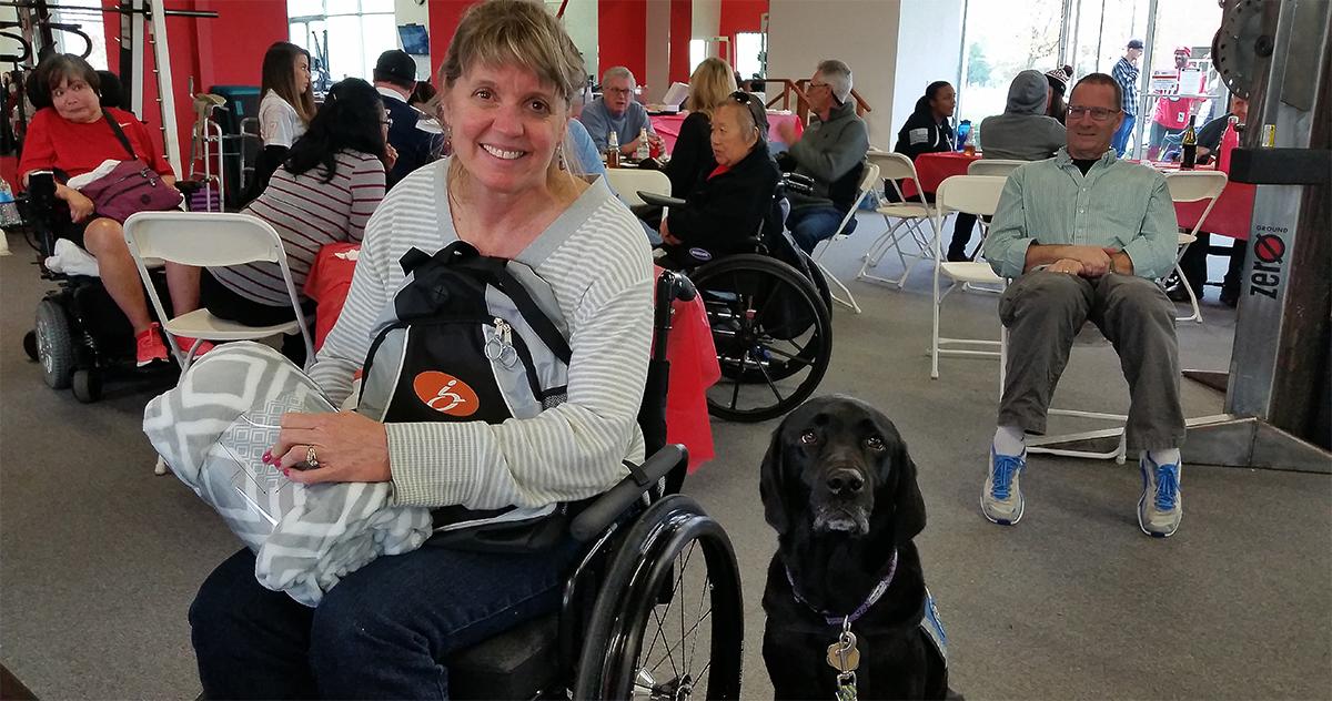 a white woman in a wheelchair holds a backpack next to her service dog