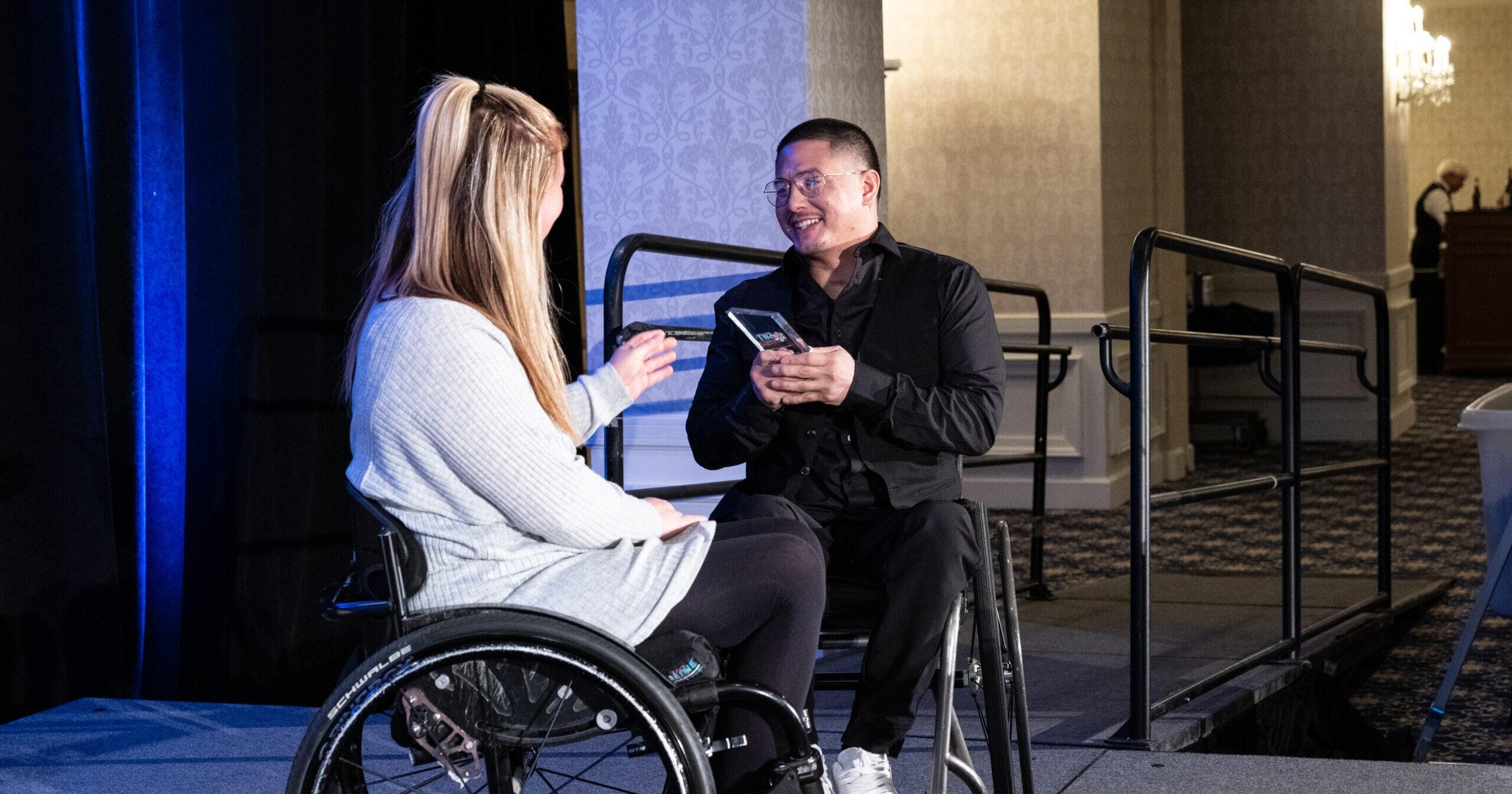 two wheelchair users on stage accepting an award trophy