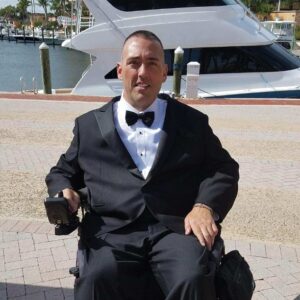 kevin mullins is a white man in a powerchair, he is wearing a suit and sitting outside.