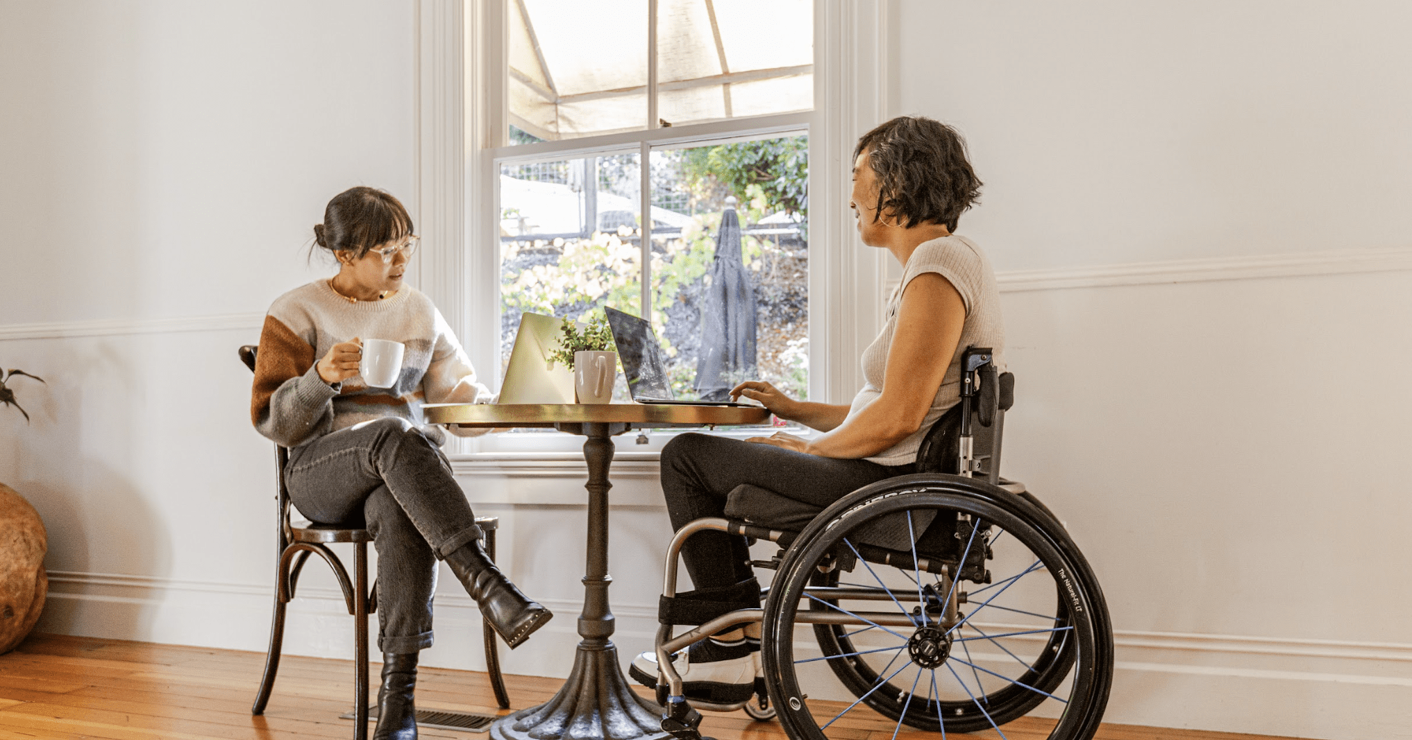 two women sitting at small table in front of a window. One is in a wheelchair