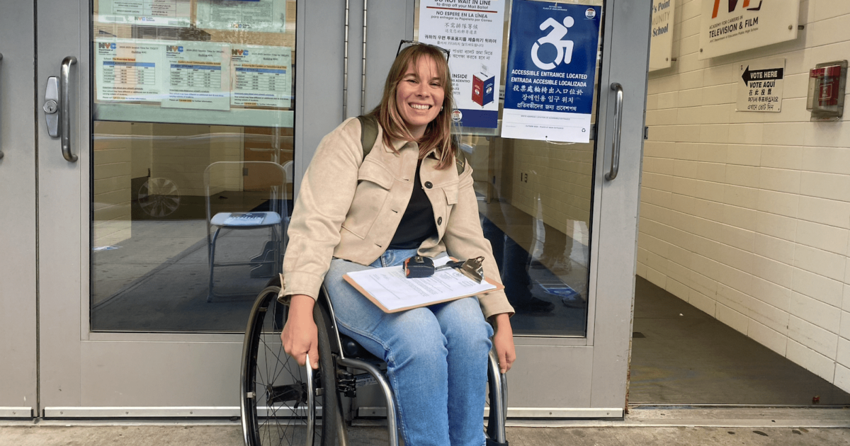 a white woman in a manual wheelchair smiles in front of a polling place