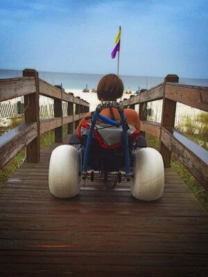 A woman faces the beach on a boardwalk in a beach wheelchair.