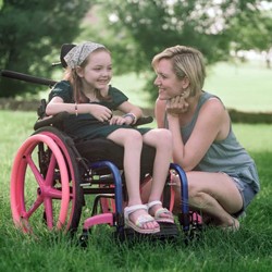 woman squatting down on the grass next to little girl in pink-wheeled wheelchair