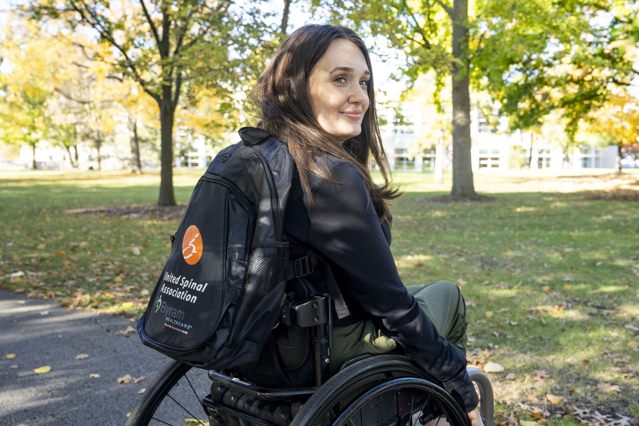 A white woman in a wheelchair sits outside in her wheelchair wearing a backpack