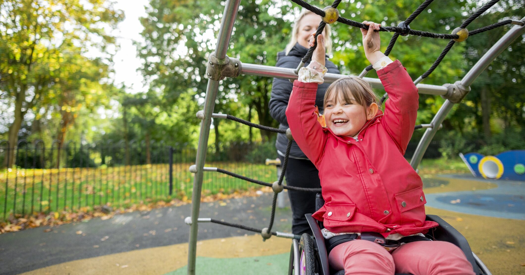A cheerful low angle medium close-up of a young girl who is a wheelchair user playing in the park on the climbing frame with her mother. They're in a playground ina. public park in Newcastle upon Tyne in the North East of England