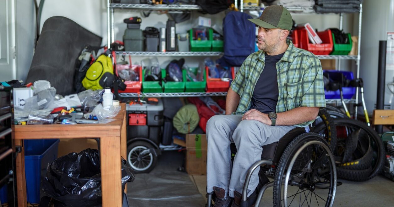 A Caucasian man with a physical disability who uses a wheelchair sits in his home garage looking at his tool bench.