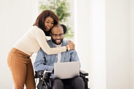 A Black woman hugs a Black man who uses a wheelchair. They are looking at his laptop together.