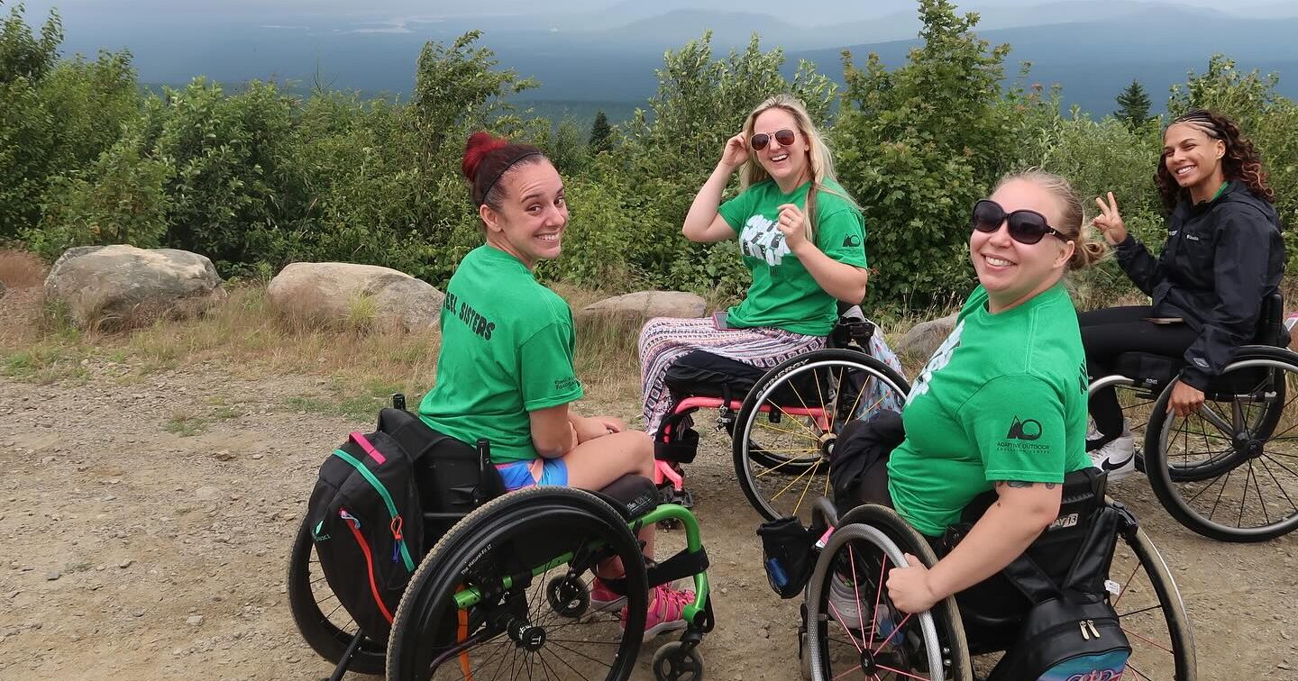 four women in wheelchairs pose outside with nature in the background