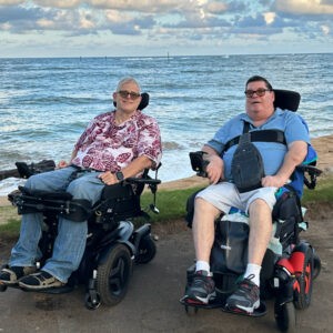 two men in powerchairs on a path by the beach with ocean in background