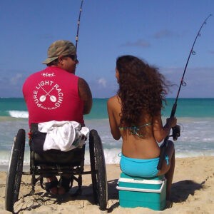 man in wheelchair fishing on the beach next to woman in bikini sitting on cooler