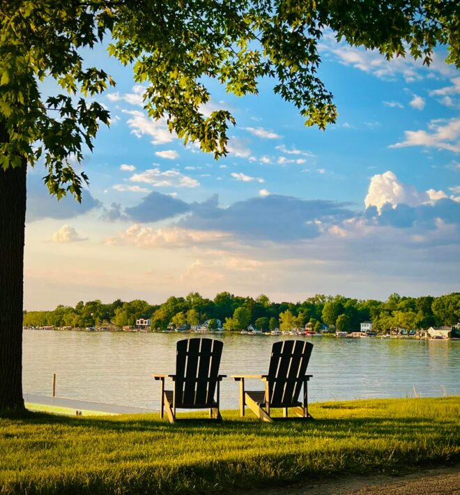 Photographic image of two empty Adirondack chairs under a tree overlooking a river with houses on the bank on the opposite bank.