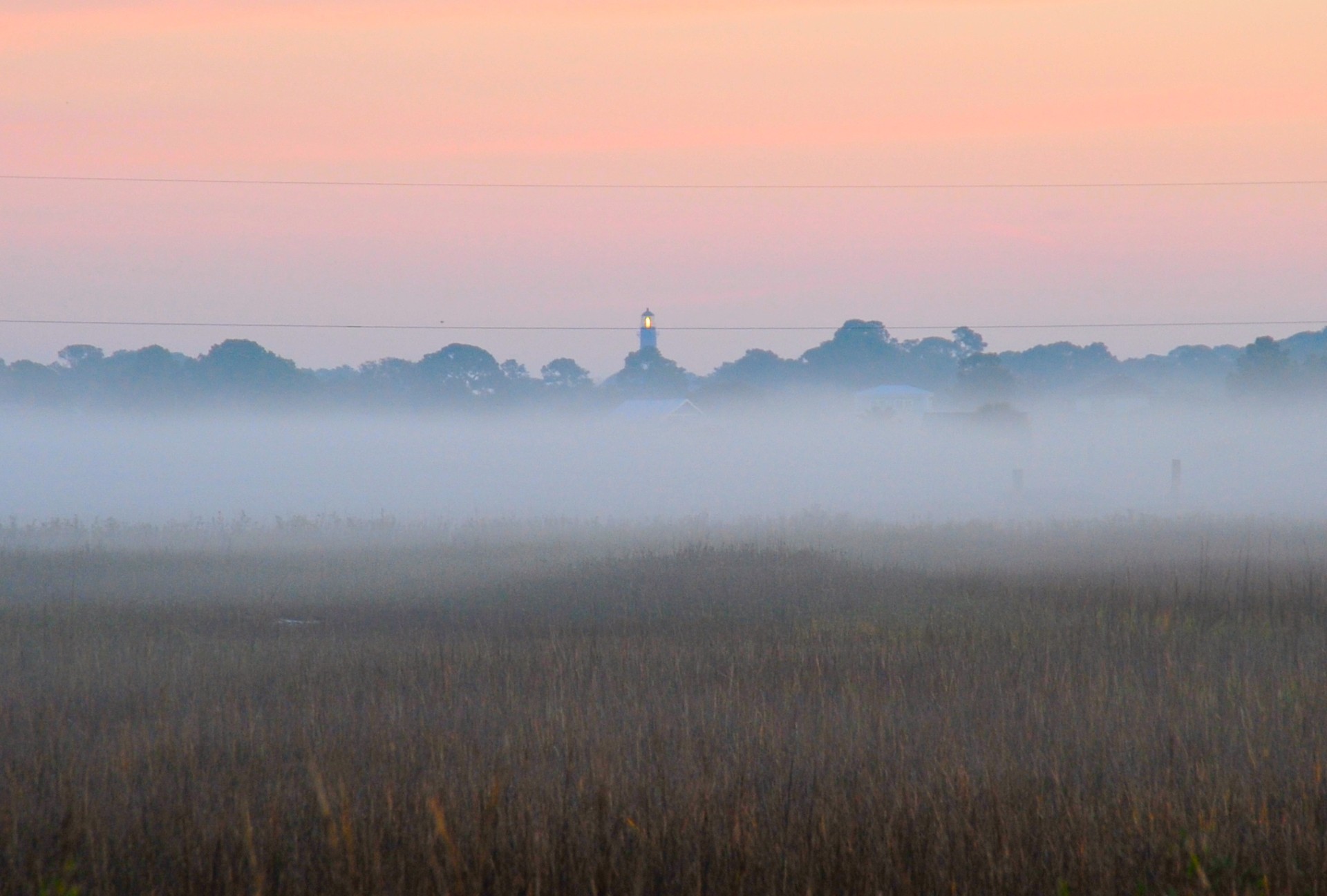 Photographic image of fog over a meadow at dusk with trees and a light beacon in the far background