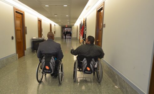 Two men using manual wheelchair roll down a hall with backpacks hanging off their chairs. 