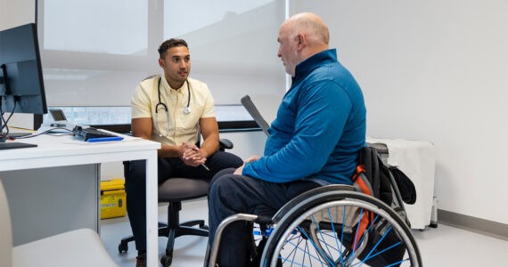 A side view of a man who is a wheelchair user meeting his doctor. His doctor is listening intently with his stethoscope around his neck.