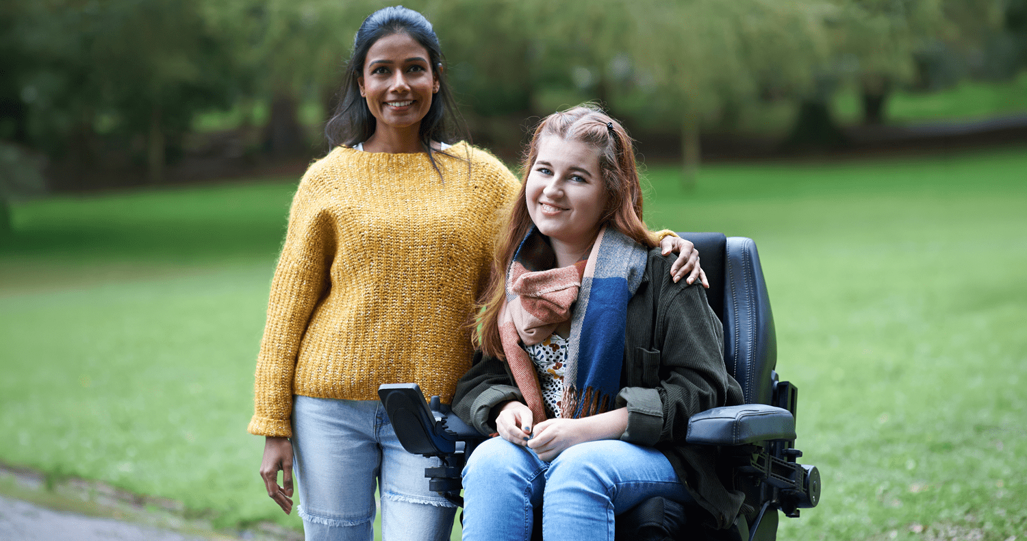 Portrait of a young woman in a power wheelchair and her friend smiling in an Autumnal park.