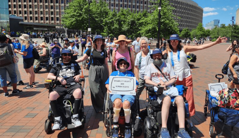 Three wheelchair users at a large crowd of people at a disability rights demonstation